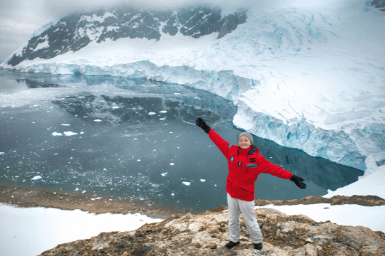 Kreuzfahrt-Gast steht im Vordergrund auf einem steinigen Felsen in der Antarktis und winkt in die Kamera. Im Hintergrund ist das Meer und die Eisberge zu erkennen.